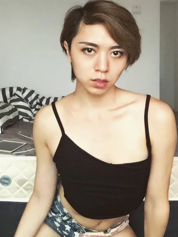 Portrait d'une personne aux cheveux courts portant un haut noir et un short, assise devant un lit dans une chambre.