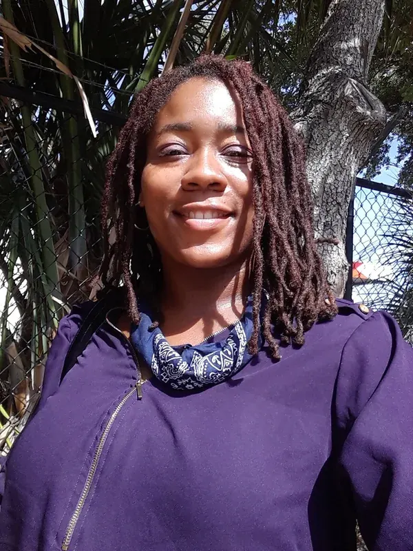 Portrait en buste d'une femme souriante avec des dreadlocks, portant une veste violette et un bandana, devant des plantes et une clôture.
