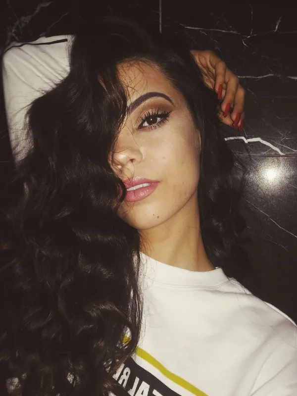 Portrait d'une femme aux cheveux noirs ondulés portant un t-shirt blanc, allongée contre un mur marbré sombre.