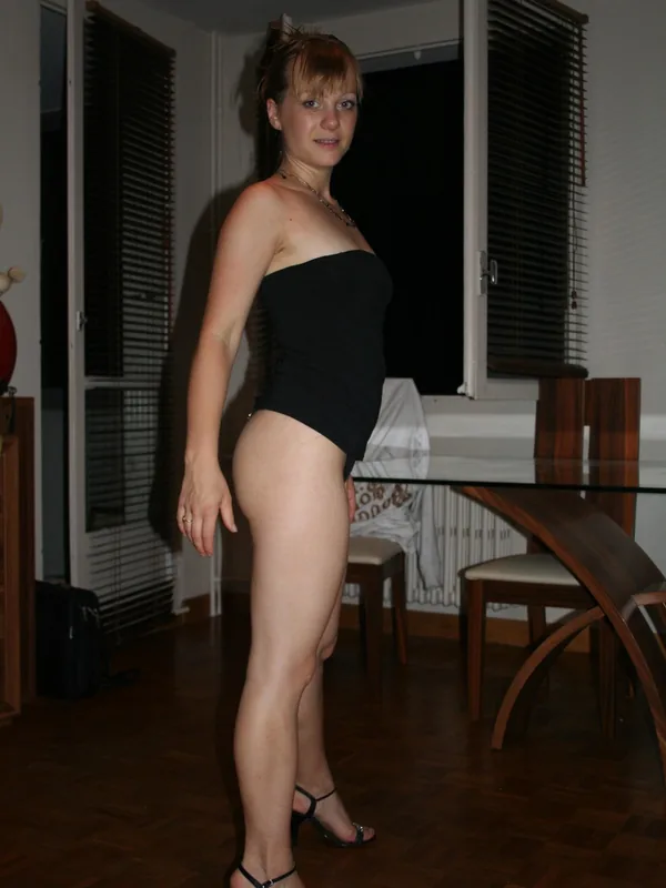 Femme debout en tenue suggestive, portant un haut noir et des talons, dans une pièce éclairée avec parquet et fenêtres aux volets fermés.