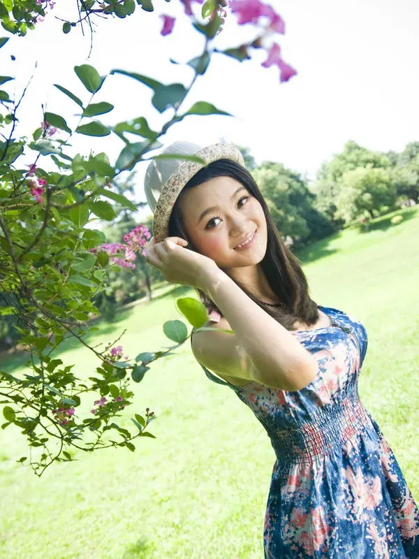 Jeune femme en robe fleurie et chapeau de paille souriant dans un parc verdoyant sous des fleurs roses.