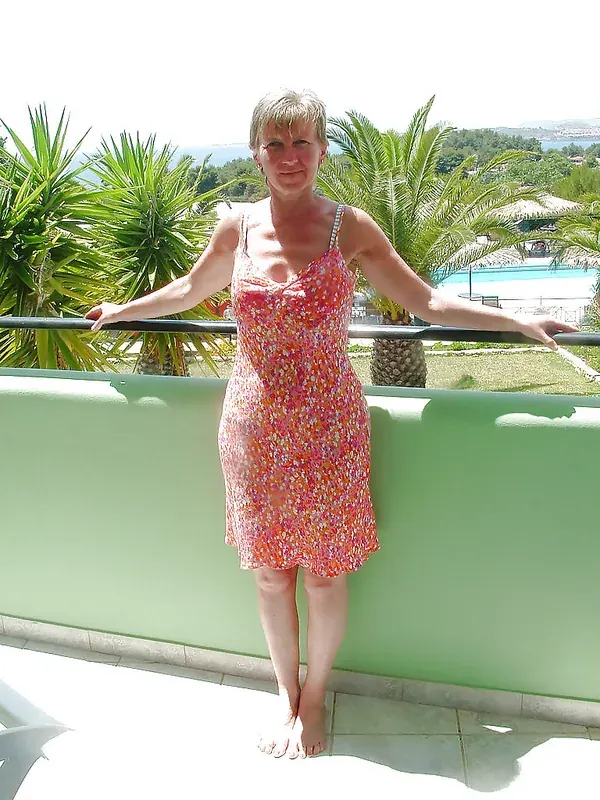 Femme en robe fleurie rose sur un balcon ensoleillé avec vue sur la mer et des palmiers.