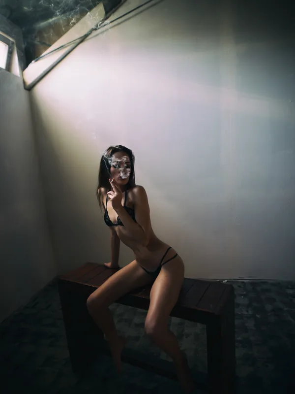 Femme en lingerie noire assise sur une table en bois, fumant, dans un espace sombre éclairé par une lumière latérale.