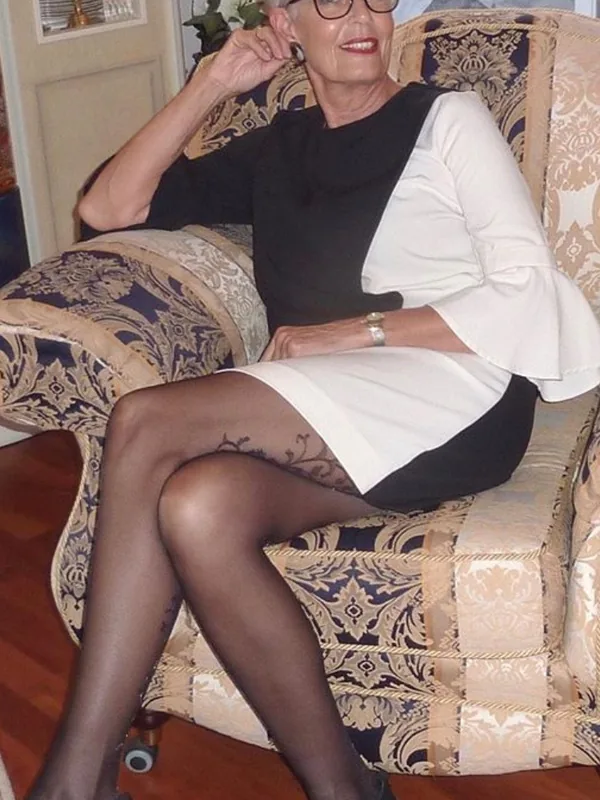 Femme aux cheveux gris portant une robe noire et blanche, assise sur un fauteuil décoratif, regardant l'objectif.