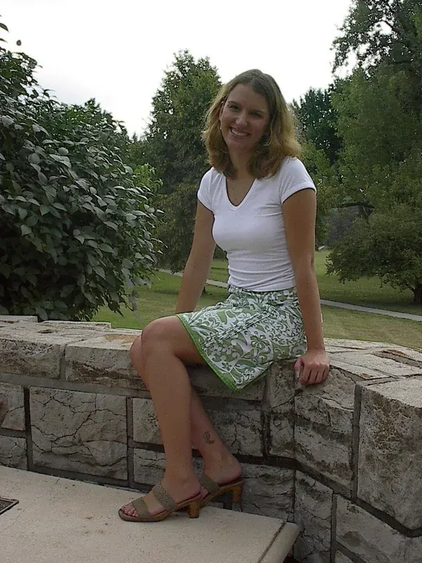 Femme souriante assise sur un mur en pierre dans un jardin verdoyant, portant un t-shirt blanc et une jupe à motifs.