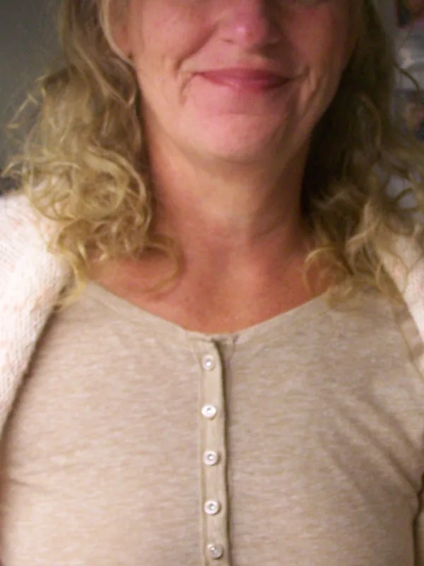 Portrait d'une femme aux cheveux blonds ondulés portant un haut beige à boutons et une veste claire, souriant légèrement.