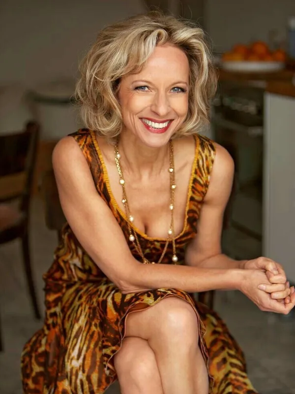 Femme aux cheveux blonds souriante, portant une robe à motifs animaliers, assise dans un intérieur lumineux.