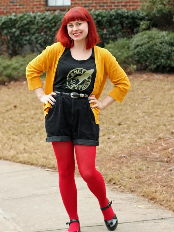 Femme aux cheveux rouges souriante, vêtue d'un cardigan jaune et de collants rouges.