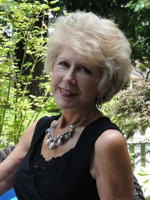 Femme aux cheveux blonds courts, souriante, portant un haut noir et un collier, assise en extérieur.